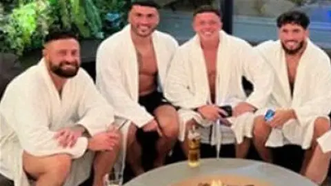 Matthew Waterworth, Regan Burnie, Paddy Nolan and Ross Boys all in white towelling dressing gowns sat round a fire pit on a spa day. They are all smiling.