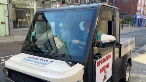Ellen Knight/BBC A small, white, two-seater vehicle with a flatbed truck at the back. On the bonnet is written 'Whitchurch Town Council' in blue letters, with 'Operation Gnome' signs taped to the side. Inside the cab of the vehicle are the two volunteers - wearing their gnome costumes.