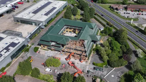 An aerial image of demolition works on a square building with a dark green roof. The building is surrounded by cranes and has a chunk taken out of its side. the roof has a number of triangular layers.