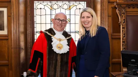 Steven Crellin, wearing a red robe with a mayoral chain, stands alongside former mayor, Natalie Byron-Teare, a woman with blonde hair dressed in a navy blue suit. Wooden panelled walls and a decorative stained-glass window can be seen in the background.