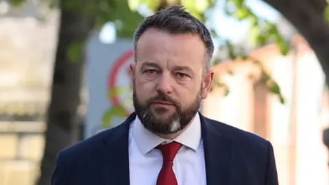 Colum Eastwood, a dark haired man with beard, wearing a navy suit, white shirt and red tie