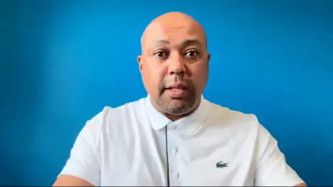 Courtney Freese A man sits in front of a blue backdrop. He is wearing a white button-up polo shirt with a Lacoste logo on it.