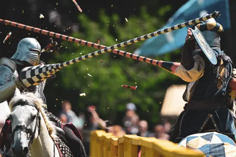 Gordy Macdonald Two people on horses with helmets jousting at an outdoor event