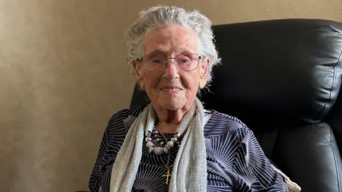 An elderly woman with short grey hair and bright eyes, smiles as she sits in a leather chair. She wears a navy shirt and grey scarf