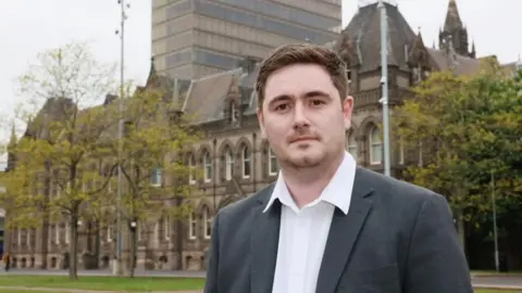 Chris Cooke looking into the camera. He's got brown, short hair and is wearing a grey blazer with white shirt. He's standing outside Middlesbrough Town Hall which is a large, gothic looking building.