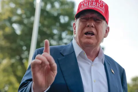 Donald Trump wearing a white shirt and navy suit jacket with no tie. He is wearing a red cap which bears the words: GULF OF AMERICA. Yet another TRUMP development.