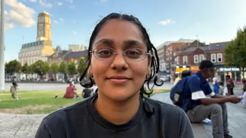Martin Heath/BBC Close-up portrait of Mansi Shah wearing glasses and a dark T-shirt, seated in a town square and looking towards the camera. Historic and modern buildings line the background, including a prominent clock tower on the left. Several people are sitting and walking around a grassy public space behind the subject. The image is taken in soft evening light with a blurred background and blue sky overhead.