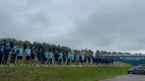 BBC  Staff and players line up on a grass verge outside Carlisle United's Brunton Park stadium