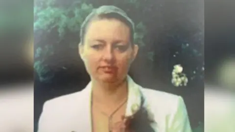 Helen Bloomfield wearing a white jacket with a flower attached to its top pocket. She has brown hair and appears to be at a wedding or similar event.