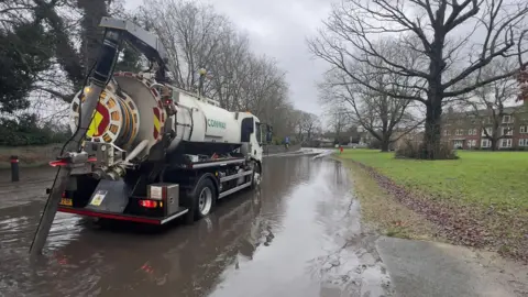 Kent County Council A white road tanker with a tube attached to the back of it. The lorry is parked on a flooded road with a patch of grass and trees to the side.