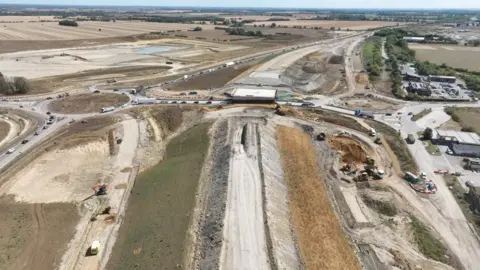 An aerial view of a large road construction project. Work is under way building up an embankment for the road heading up the middle of the picture, including the beginnings of a bridge over an existing road. Traffic is using a roundabout on the left.
