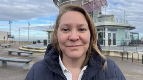 Louise Hobson/BBC Anna Turley is stood at Redcar seafront where she hopes to build a new pier for the town. She is smiling at the camera and wearing a navy coat over a white shirt.