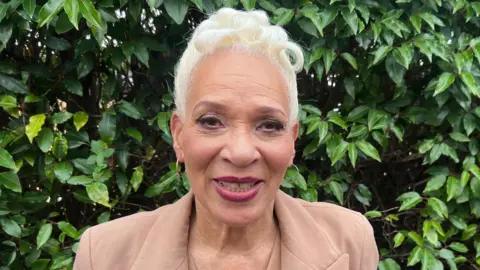 Lily-May Symonds/BBC Lena Barnes is in the middle of the frame. She has short white hair and is wearing a light pink blazer. She is stood in front of a hedge.