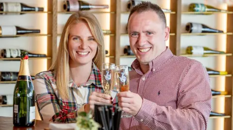 National Lottery/PA Wire Luke Harris (on the right, a man with short brown hair and smiling looking into the camera, wearing a red and white chequered shirt) and Alison Coke (on the left, a woman with blonde hair looking directly into the camera and smiling, wearing a patterned dress consisting of white, green and red) each holding a glass of champagne and celebrating their win