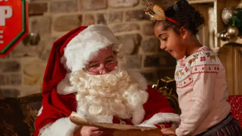 A man dressed as Father Christmas in traditional white-trimmed red clothes is leaning over a book, while a child of around eight years old, wearing a Christmas decoration headband stands by him also leaning over the book.