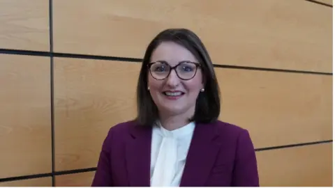 Department of Health Emma with glasses and shoulder length hair. She is wearing a purple blazer and a white high neck top. She has pearl earrings and red lipstick on. 