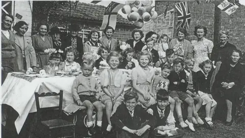 A black and white photo showing a group of people, mainly children, in 1950s clothing, sat around a table smiling at the camera, with union flags hanging in the background.