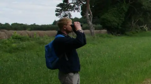 RSPB Tim Cowan is pictured in a grass field looking through binoculars for stone-curlew birds. He has short blond hair and is wearing a black jumper with grey trousers. He has a blue backpack on.