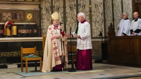 Church of Saint Mary of the Isle John Sherrington wears an ornately decorated cream and gold mitre and a white and red robe in a cathedral. A men in a maroon and white robe stands beside him.