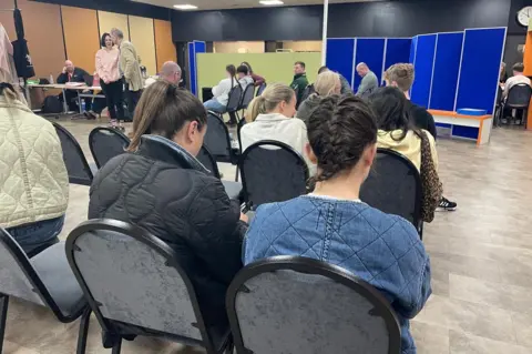 Stephen Huntley/BBC Young people, including two women nearest the camera, are sitting on chairs with their backs turned. They are inside a hall or large room.