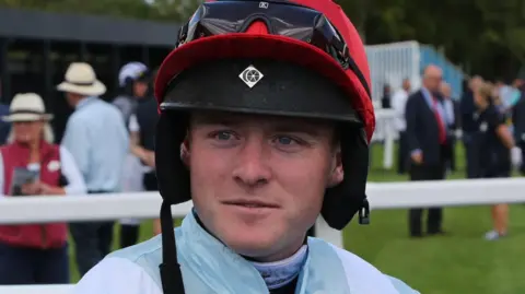 Oliver Boyden The photo shows a person dressed in full racing attire standing outdoors at what appears to be a horse‑racing venue. The individual is wearing a red racing helmet with protective goggles resting on the front, a pale blue and white racing jacket, and white riding trousers secured with a belt. A black glove is visible on one hand, and the person is holding a small object that looks like part of their racing equipment.