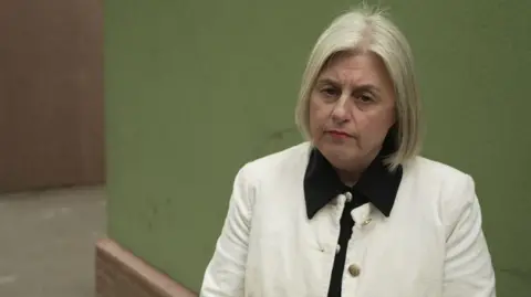 Michelle Walsh has short blonde hair, is wearing a white jacket, and is pictured in front of a green wall. 