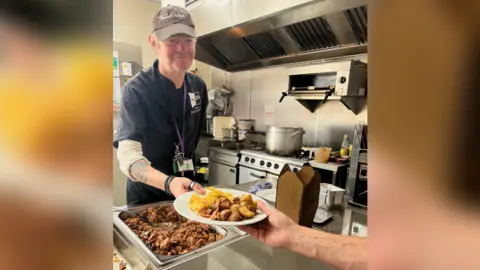 Cheltenham Open Door A man wearing chef uniform stands in a commercial kitchen. He smiles as he hands someone out of shot a plate of food.