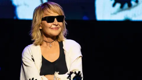 Getty Images Lulu, who has blonde, shoulder-length hair, is dressed in a white shirt and black top, and is wearing sunglasses. She has her arms folded and is stood in front of a black background.