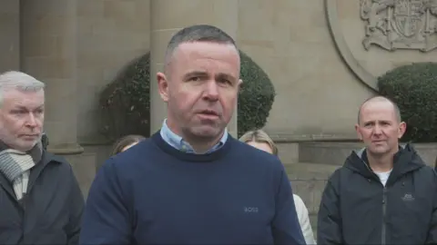 A man with short dark hair and wearing a blue jumper reads a statement from his phone as several family members stand behind him outside the court building