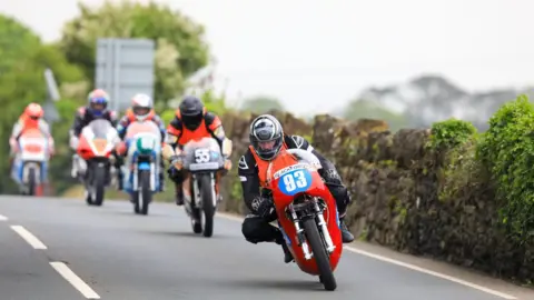 CALLUM STALEY A line of motorcycles heading over the brow of the hill. The front bike is red and has the number 93 on the front. There is a stone wall to the left.