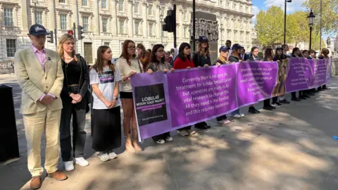 Chrissie Reidy/BBC A group of people standing behind a long purple banner, on a footpath, near a government building.