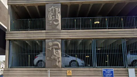 Google Exterior shot showing two floors of a concrete multi-storey car park showing abstract designs in relief on the pillars and blue metal railings in between. Two cars are visible parked inside on the lower floor.