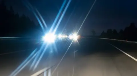 A shot taken at night on a dual carriageway. The carriageway on the right is empty. On the other carriageway, the bright lights of car headlights are flaring in the lens of the camera as they drive towards it. The silhouettes of trees can be seen on either side of the road.