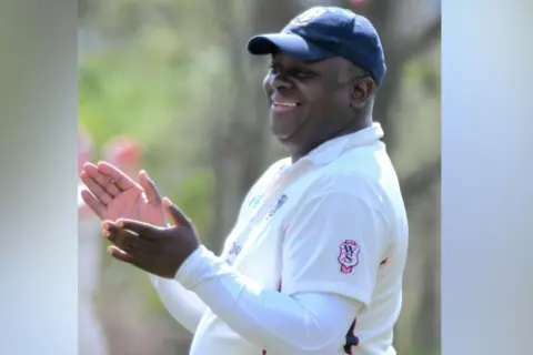 David Randall Foundation Norman Case stands on a cricket pitch, wearing cricket whites. He is also wearing a navy blue baseball cap. He is smiling and clapping his hands