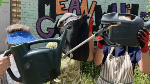 Three women standing in front of a brick wall. The woman in the middle is holding a black bag over her face and those to either side of her are holding plastic watering cans over their faces.