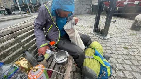 Nick Brooke Nick Brooke sits on a park bench next to his his belongings, including a camping stove, water bottles and a tin of instant coffee. At his feet is a blue rucksack with a yellow sleeping bag trapped to it. He is wearing a grey hiking coat with another blue waterproof coat underneath with the hood up and he is peering down into a white bag.