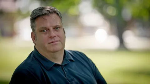 BBC Steven is wearing a blue shirt and is sitting on a bench outside. He is in focus but the background, which resembles grass and trees, is fully out of focus. Steven is looking directly into the camera with a straight face.
