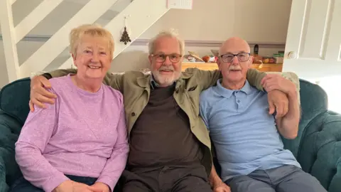 Photo of Kjell-Åke with his arms around Kath Francis who is on his right side and Ray Francis who is on his left side. Kjell-Åke is wearing a black jumper and black trousers, grey collar shirt and a green jacket. He has grey hair and a grey beard and he's wearing glasses. Kath has short blonde hair and a pink jumper and Ray is bald with glasses and is wearing a blue polo tshirt and grey trousers. Their sat on a dark green sofa in a living room.