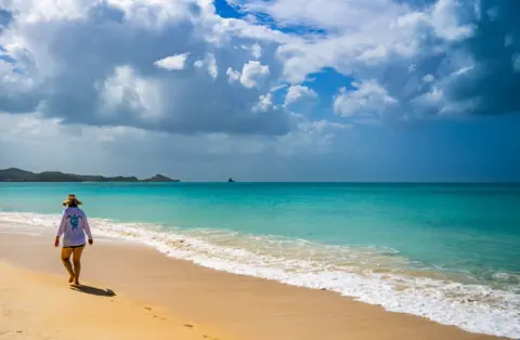 Carlos Gomez Llata ​​Seorang wanita di sunhat berjalan di sepanjang pantai di hari yang cerah. Gambar menunjukkan dia berjalan menjauh dari kamera. Lautnya tenang dan pirus dan langit berwarna biru dan mendung.