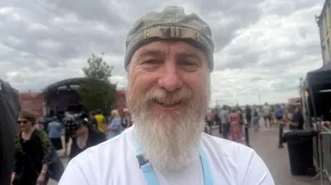 Mark Page - a man in a grey cap with a long grey beard and moustache is smiling into the camera lens. He is wearing a white top, blue lanyard and you can see a stage and people dotted about on a street behind him.