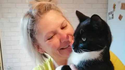 Maccy G Pets A blonde woman wearing a yellow top holds a black and white cat