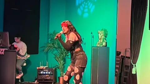 SIMON DEDMAN/BBC A drag artist is singing on stage. She has long black hair with a red fringe. She is holding a microphone and is dressed all in black with holes in her tights.