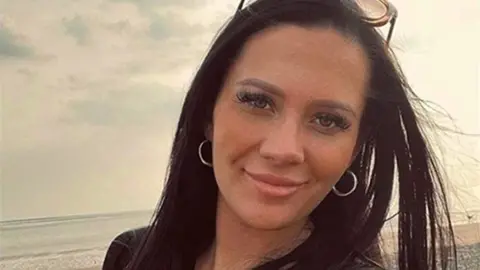 Kiena Dawes, with long black hair and silver hoop earrings, smiles as she is photographed on a beach.