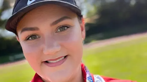 A young woman, who wears a black sports cap and a red t shirt, smiles from a playing field.
