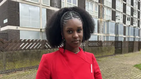 A woman wearing a red jacket stands in front of a housing block with numerous boarded up windows