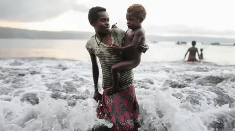 A small child is carried by an adult amid waves on Pacific island of Vanuatu, which is threatened by rising seas