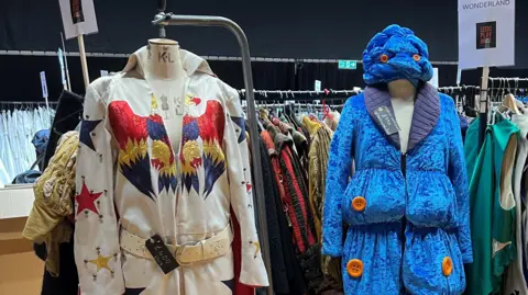 Amanda Harper/BBC Two costumes on mannequins. To the left is an Elvis-style jumpsuit in white with gold, red and blue detailing. On the right is a blue velvet coat and hat with bulges.