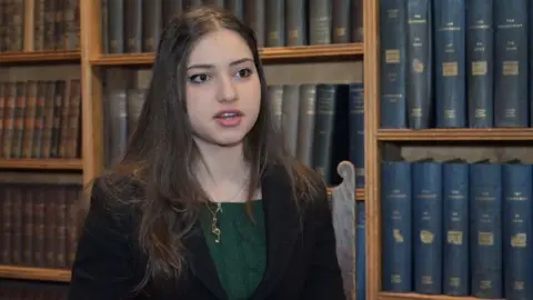 Arwa Elrayess is standing in front of old bookshelves. She has long dark hair and is wearing a dark blazer over a green jumper.