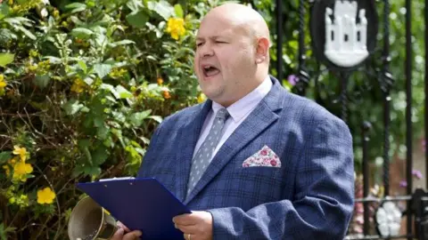 Rich Whincup wearing a suit and holding a clipboard while shouting in the town