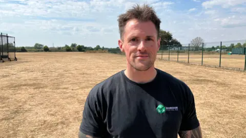 Andrew Briggs stands on a dried out sports pitch. The grass is brown and yellow after weeks of drought. He is wearing a black T shirt with a small green symbol on it, and has short dark brown hair
